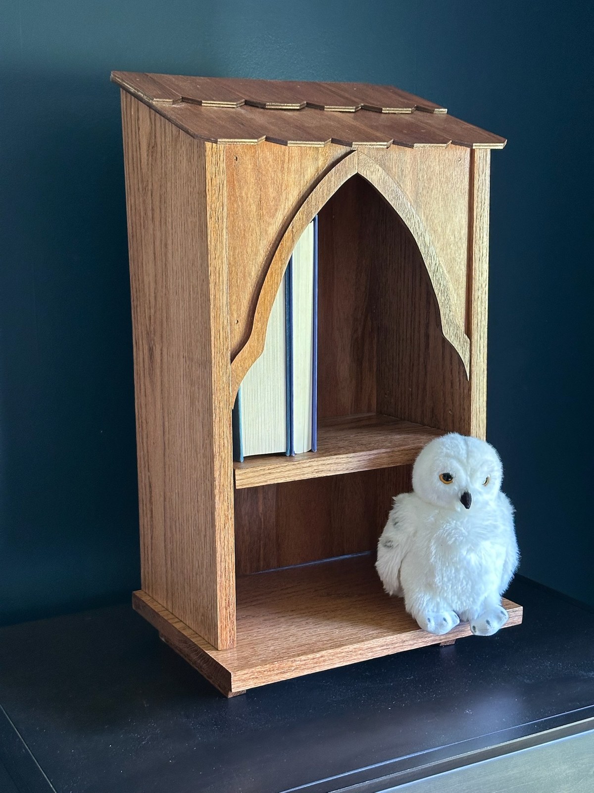 Angled view of a wooden Owlery bookshelf with a pointed arch, layered roof details, books, and a stuffed white owl