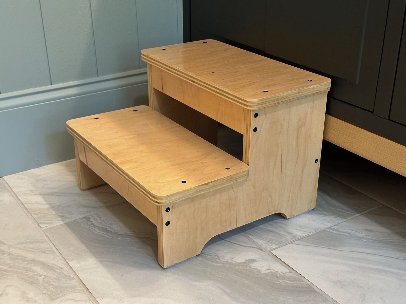 Angled view of the completed wooden two-step stool, showing its exposed plywood edges, rounded treads, and side panels